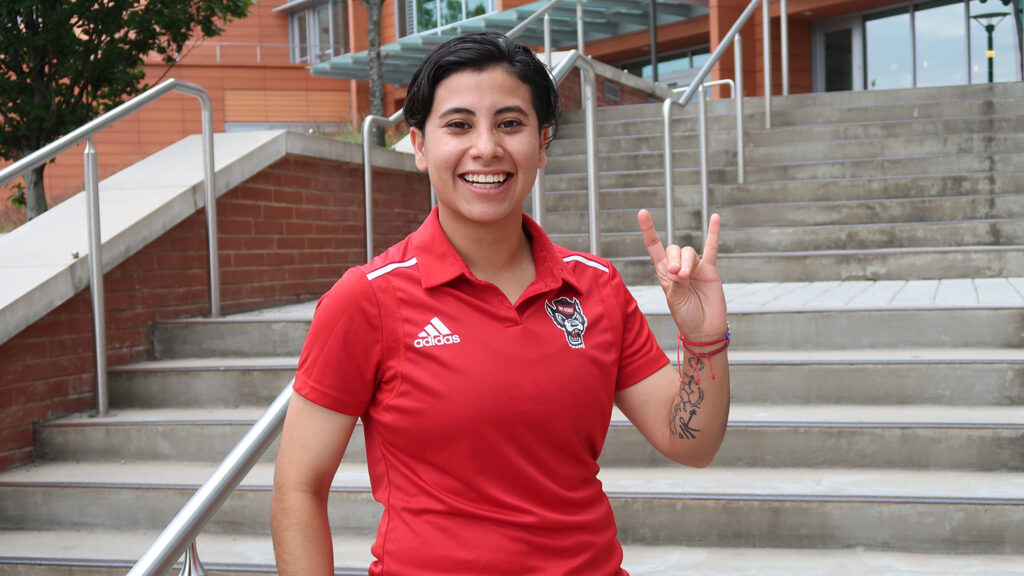 Daisy Aguilar Aguilar flashes the NC State wolf hand sign while standing at the foot of the stairs at Talley Student Union.