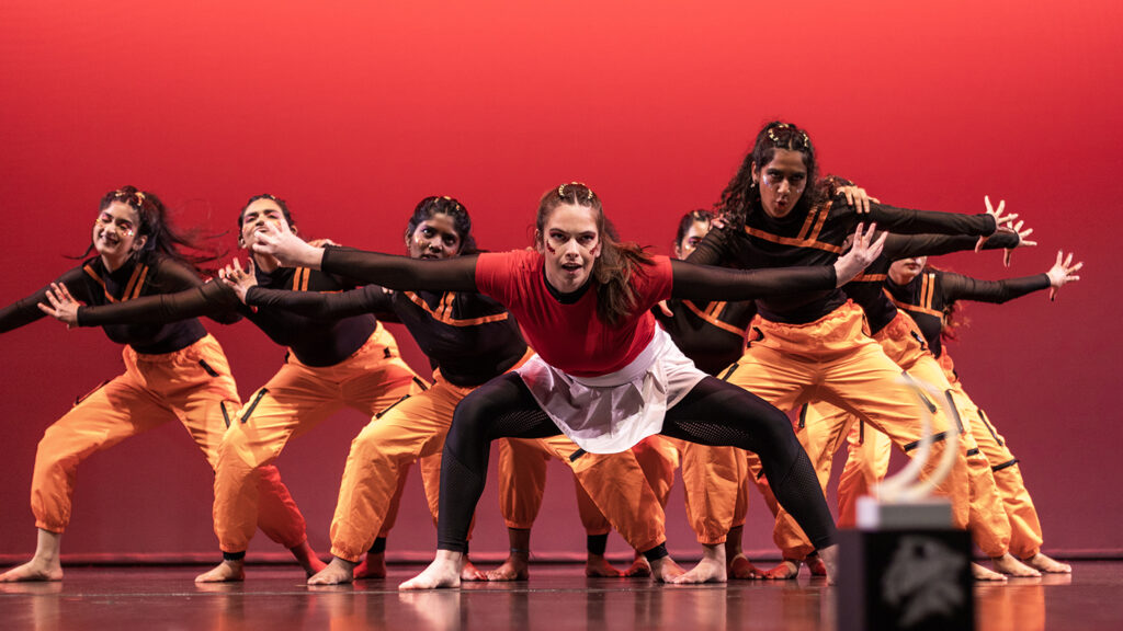 Kaia Spero leads a dance troupe on stage with a red background.