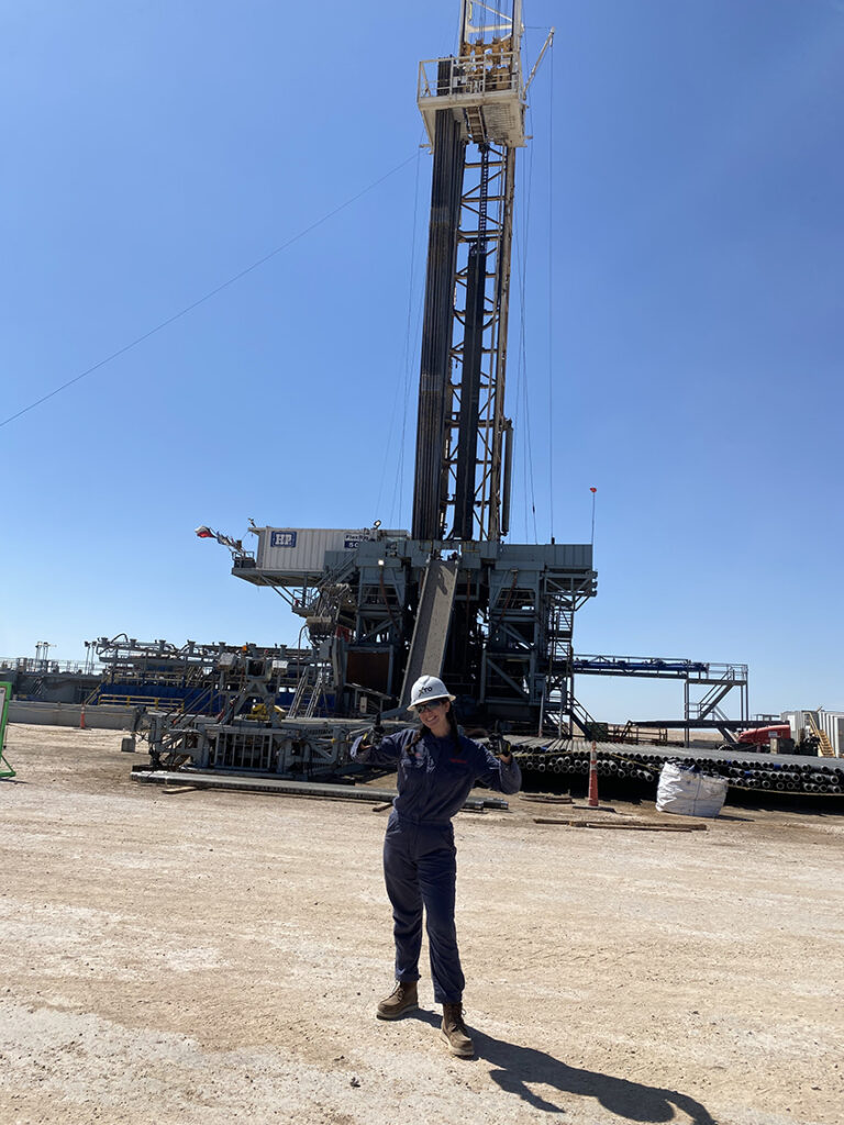 Kaia Spero in front of a drilling rig. She is wearing a white hardhat and dark coveralls.