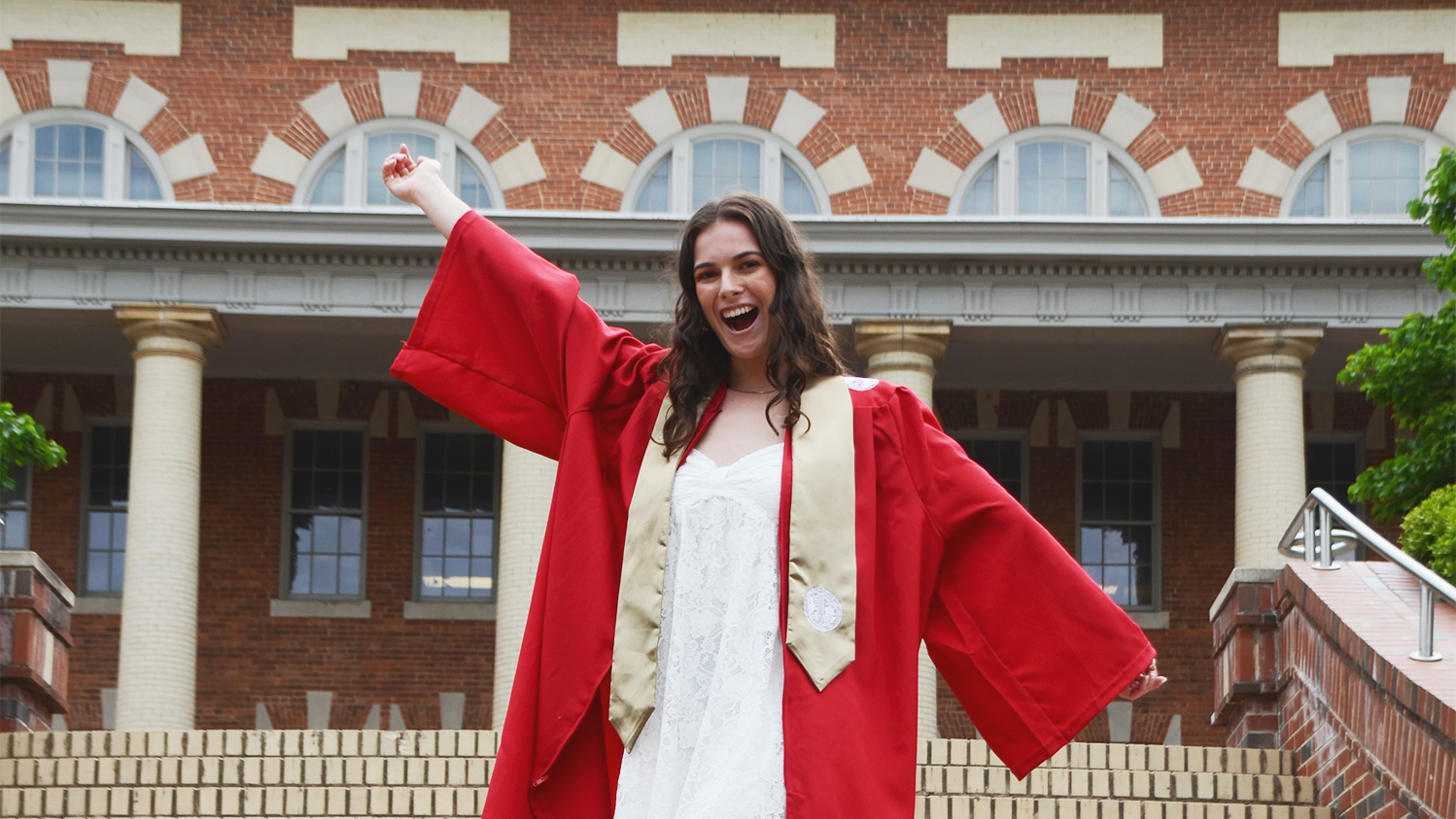 Kaia Spero, dressed in a red graduation gown and white dress, strikes a celebratory pose in front of brick building with columns.