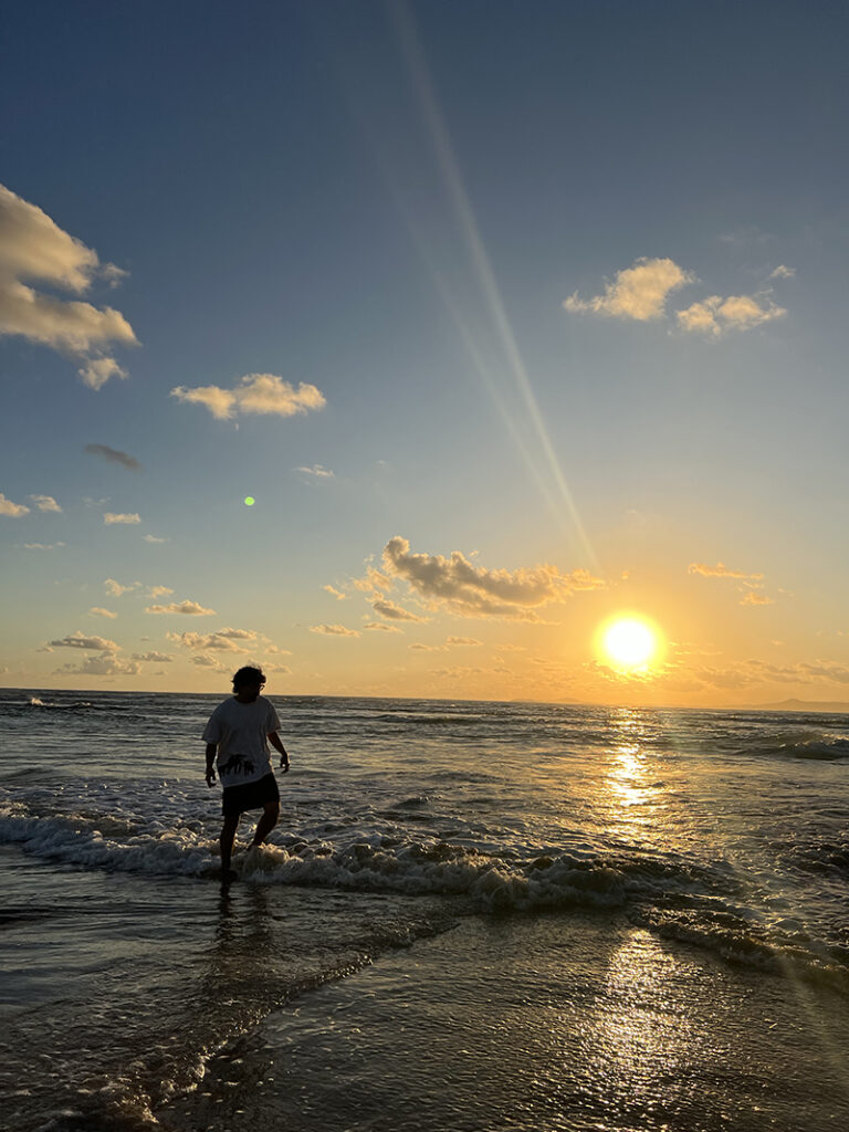 Manny Valbuena, in shadow silhouette, stands at edge of beach and ocean. A warm yellow sunset is in the background to the right. A dark blue to black sky is above punctuated with a few small gray and white clouds.
