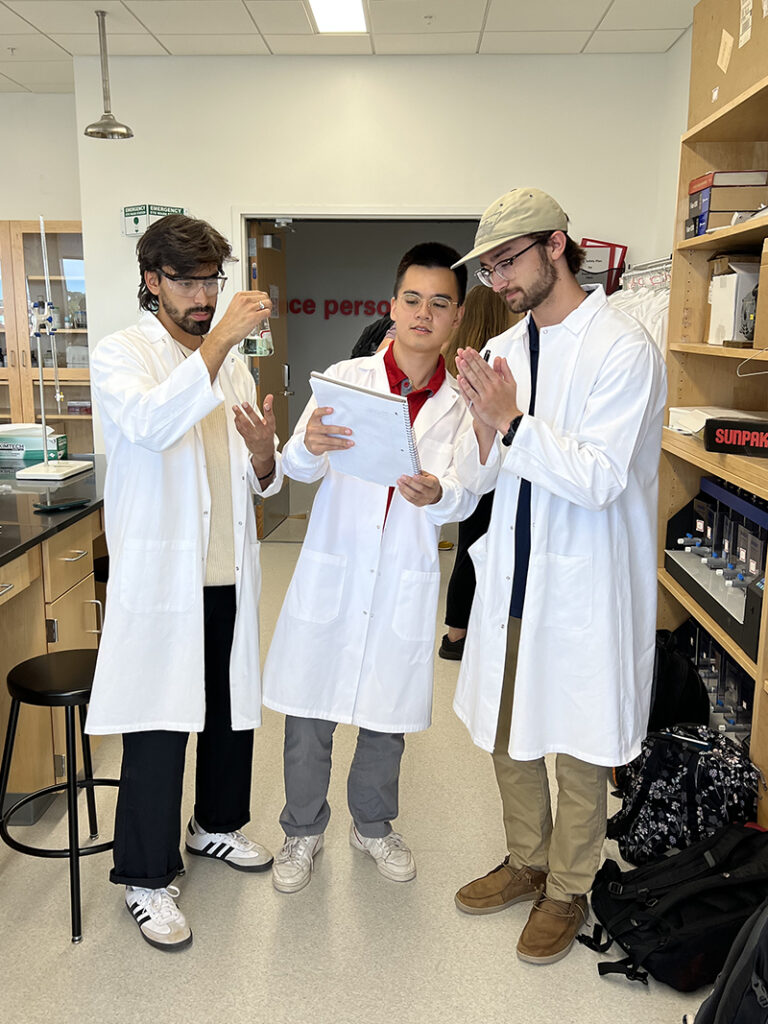 Manny Valbuena, left, dressed in a white lab coat and PPE works with two other students dressed in matching PPE.