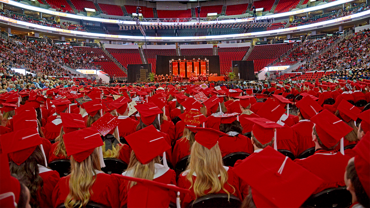 Graduating students dressed in red caps and gowns sit together on the floor of the PNC Arena during a commencement ceremony. The mid and upper seating decks and event stage are visible in the background.