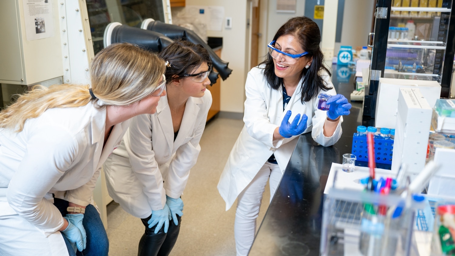 In a lab wearing a lab coat, gloves and goggles, Januka Budhathoki-Uprety (right) explains a concept to Ashley Lamb and Hannah Dewey.
