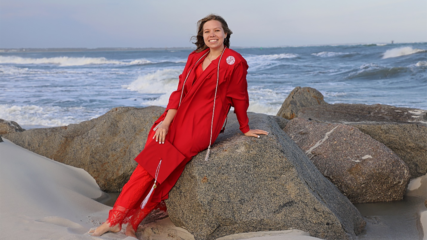 Savannah Tabor, dressed in NC State red graduation gown, leans against large boulders on Fort Macon Beach. The Atlantic Ocean and waves can be seen behind her.