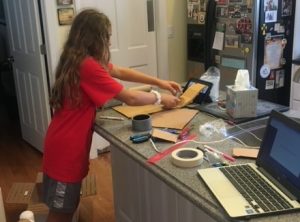 Elementary student designing a solution to a challenge using cardboard, tape, and other supplies. A laptop computer is next to her. 